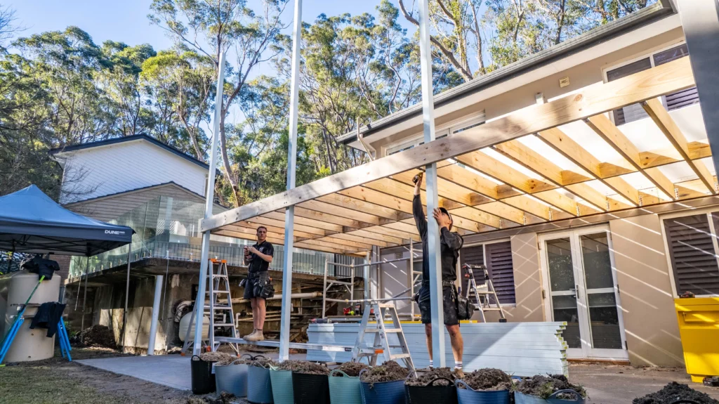 Builders framing a timber pergola extension in the backyard of a modern residential house surrounded by trees.